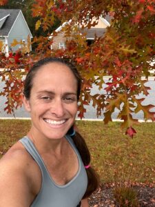 A female runner smiling while standing next to a tree with pretty fall colored leaves on it.